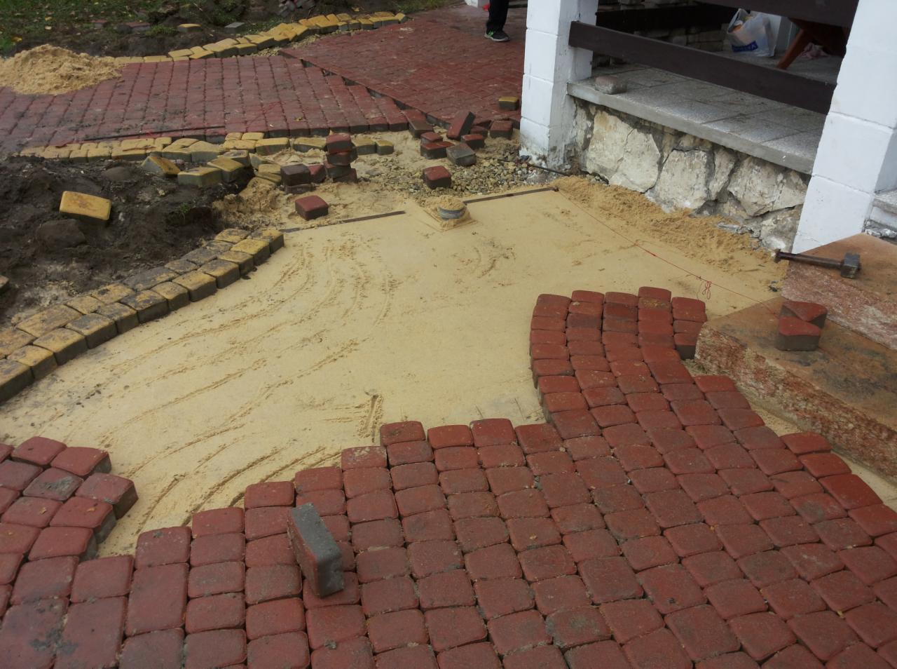 Układanie czerwonej kostki brukowej na podsypce piaskowej. Częściowo ułożony wzór, widoczne narzędzia i fragment istniejącego chodnika. Prace brukarskie w toku.