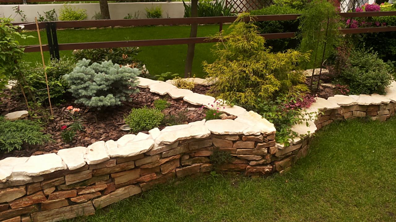 Ogród z kamiennym murkiem oddzielającym rabatę z korą, iglakami i kwiatami od trawnika. W tle drewniane ogrodzenie i trawnik.