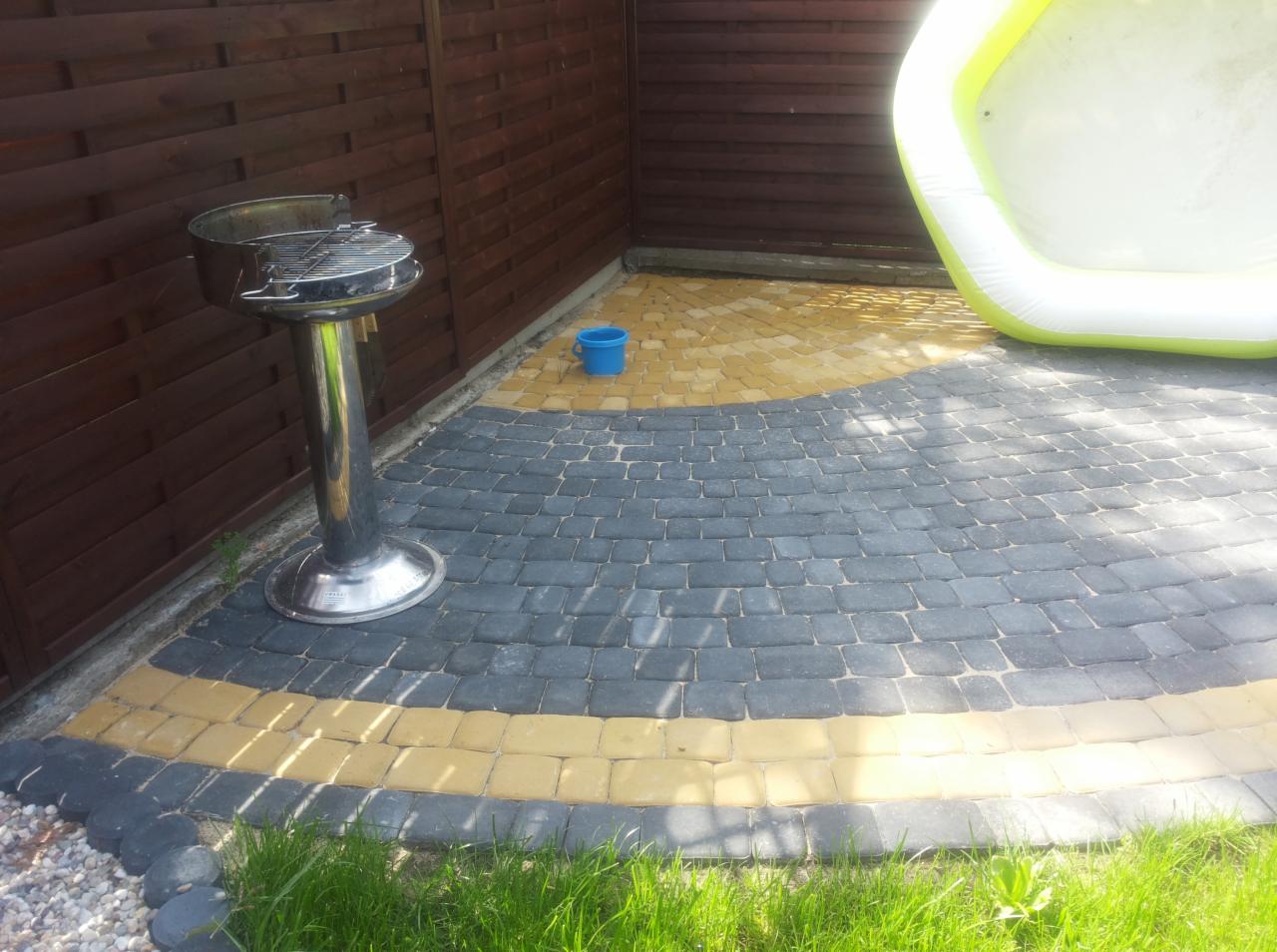Ułożona kostka brukowa w odcieniach szarości i żółci, tworząca wzór na podwórku. Widoczny grill, mały basen i drewniane ogrodzenie w tle.