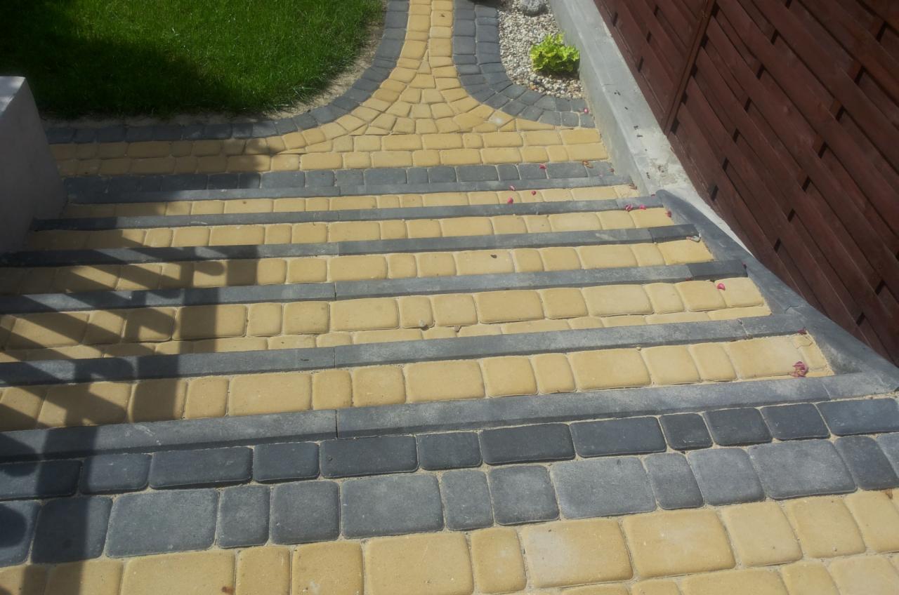 Ułożone schody z kostki brukowej w odcieniach szarości i beżu, prowadzące do drewnianego ogrodzenia. Widoczne elementy zieleni i drobne kwiaty.