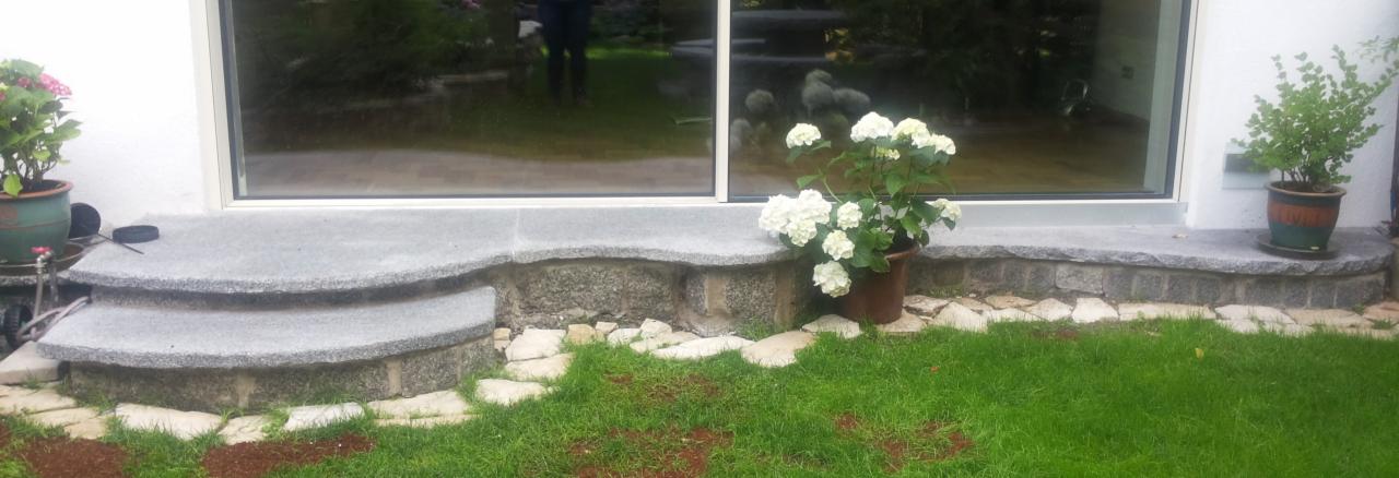 Granitowe schody wejściowe do budynku, z doniczkami z kwiatami po bokach. Widoczna trawa i fragment elewacji. Kamienna podstawa schodów.