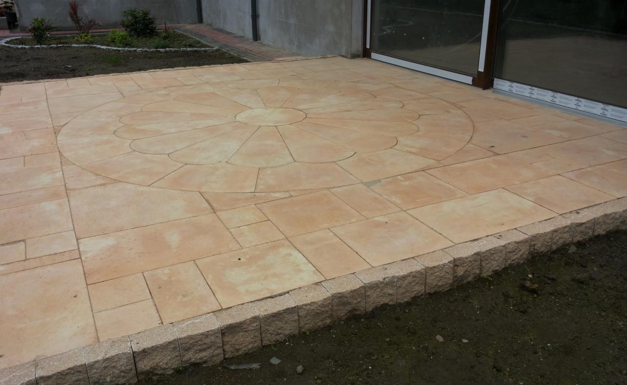 Beżowy taras z kostki brukowej z dekoracyjnym wzorem koła i obrzeżem z kamienia, przylegający do budynku z oknami tarasowymi. Widoczny fragment rabaty.