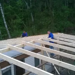 Montaż drewnianej konstrukcji dachu przez dwóch pracowników w Pilawie. Widok na szkielet z desek na tle zieleni drzew.