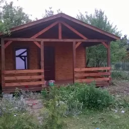 Drewniana altana z oknem i zadaszeniem, otoczona zielenią. Na pierwszym planie roślinność i ścieżka, w tle widoczny drugi budynek. Całość w naturalnej kolorystyce.