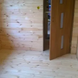 Jasne, drewniane wnętrze z boazerią, widoczne podłogi i ściany. Otwarta, brązowa, nowoczesna  stolarka drzwiowa z przeszkleniami. W tle widoczne wyposażenie pomieszczenia.