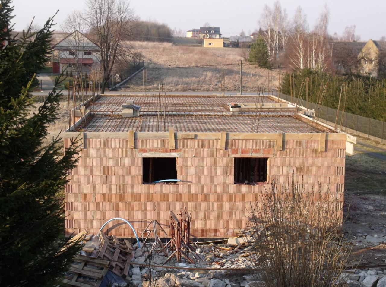 Surowy budynek z czerwonej cegły z otworami okiennymi i zbrojoną płytą stropową w wiejskim otoczeniu. Widoczne rusztowania i materiały budowlane.