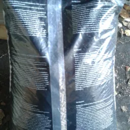 Worek z pelletem drzewnym w czarnym, foliowym opakowaniu, z widocznym tekstem informacyjnym w kilku językach, leżący na ziemi.