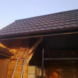 Brązowy dach z blachy trapezowej na drewnianej konstrukcji. Widoczne rynny i drabina oparta o ścianę budynku. Ujęcie z dołu, w słoneczny dzień.