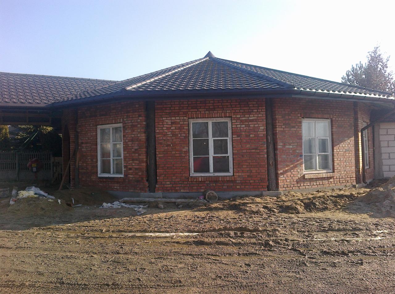 Okrągły budynek z cegły z ciemnym dachem z dachówki ceramicznej i rynnami. Widoczne okna z białymi ramami i ziemia przed budynkiem. Nietypowy kształt dachu.