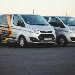 Dwa srebrne busy Ford Transit zaparkowane obok siebie; jeden z brandingiem firmy transportowej Martex z informacją o licencjonowanym przewozie osób do Niemiec, Holandii, Luksemburga i Szwajcarii.