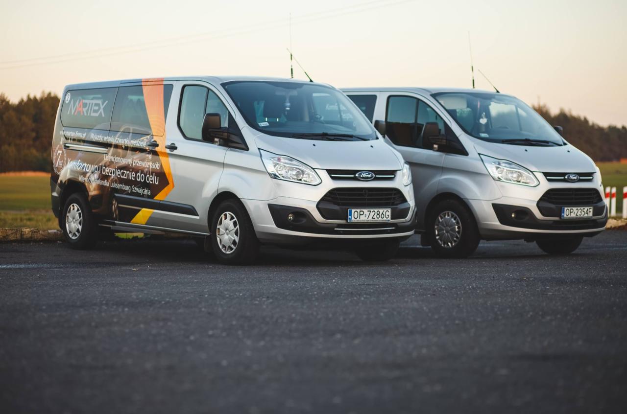 Dwa srebrne busy Ford Transit zaparkowane obok siebie; jeden z brandingiem firmy transportowej Martex z informacją o licencjonowanym przewozie osób do Niemiec, Holandii, Luksemburga i Szwajcarii.