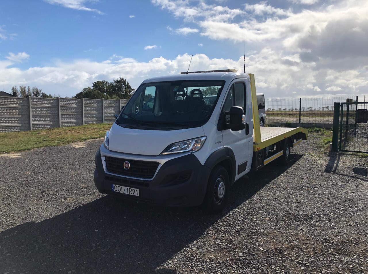 Biała laweta marki Fiat Ducato na żwirowym placu, widoczna tablica rejestracyjna D0L 1R91, ogrodzenie z betonowych płyt w tle.