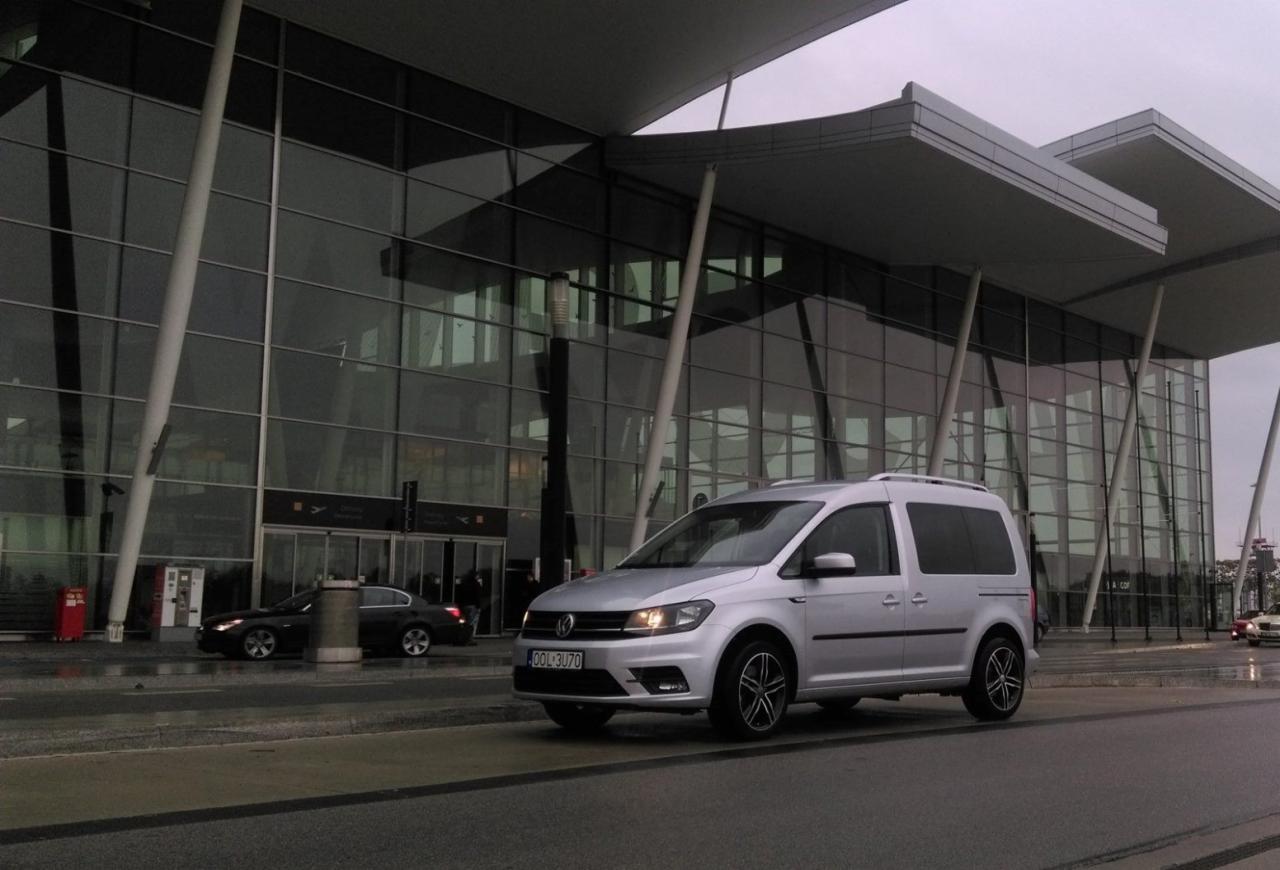 Srebrny Volkswagen Caddy z relingami dachowymi zaparkowany przed nowoczesnym budynkiem lotniska z przeszkloną fasadą w pochmurny dzień.