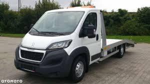 Biała laweta marki Peugeot z pomarańczowym światłem ostrzegawczym na dachu, zaparkowana na szarym parkingu z widocznymi drzewami w tle.