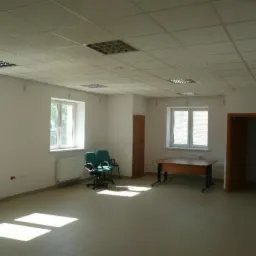 Jasne, przestronne pomieszczenie biurowe z podwieszanym sufitem, oknami i prostym umeblowaniem. Widoczne dwa krzesła obrotowe i biurko. Świeże, białe ściany.