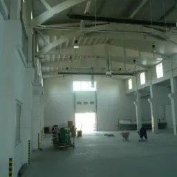 Wnętrze hali produkcyjnej po remoncie, świeżo pomalowane ściany i sufit na biało, widoczne okna, instalacje pod sufitem i sprzęt budowlany.