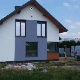 Dwukolorowa elewacja domu jednorodzinnego z szarą częścią pomalowaną na gładko i białą z wyraźną fakturą, widoczny firmowy bus z logo na tle szklarni.