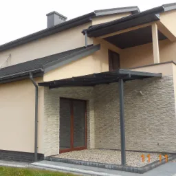 Dwupiętrowy dom z beżową elewacją i fragmentem ściany wykończonym kamieniem elewacyjnym, zadaszenie tarasu wsparte na metalowych słupach.