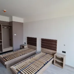 Pokój hotelowy z dwoma łóżkami z drewnianymi stelażami i brązowymi zagłówkami, szafkami nocnymi oraz dużą szafą wnękową z półkami i drążkiem na ubrania, świeżo po remoncie, widoczne ślady po...