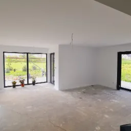 Wnętrze domu w trakcie wykańczania: świeżo pomalowane, białe ściany, surowy beton na podłodze, duże okna z widokiem na zieleń, wystające kable oświetleniowe.