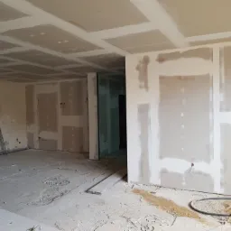 Wnętrze w trakcie prac wykończeniowych: ściany i sufit pokryte płytami gipsowo-kartonowymi, widoczne łączenia szpachlowane, na podłodze gruz i piasek, drabina oparta o ścianę w tle.