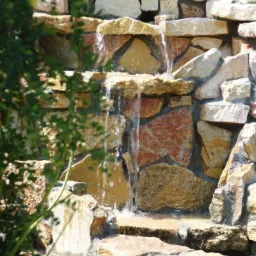 Ogród skalny z kaskadą wodną. Naturalny kamień w odcieniach beżu i brązu tworzy malowniczy wodospad, otoczony zielenią. Ujęcie z bliska.