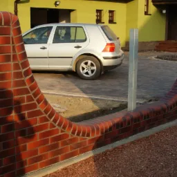 Ceglany, falisty murek ogrodzeniowy z metalowym słupkiem na tle domu i kostki brukowej. Widoczny cień osoby na murku. Ujęcie z dołu.