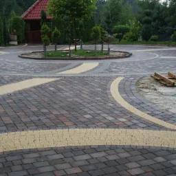 Rozległy, okrągły podjazd z kostki brukowej w odcieniach brązu i beżu, z centralnym klombem i elementami krajobrazu w tle. Widoczne ślady prac budowlanych.