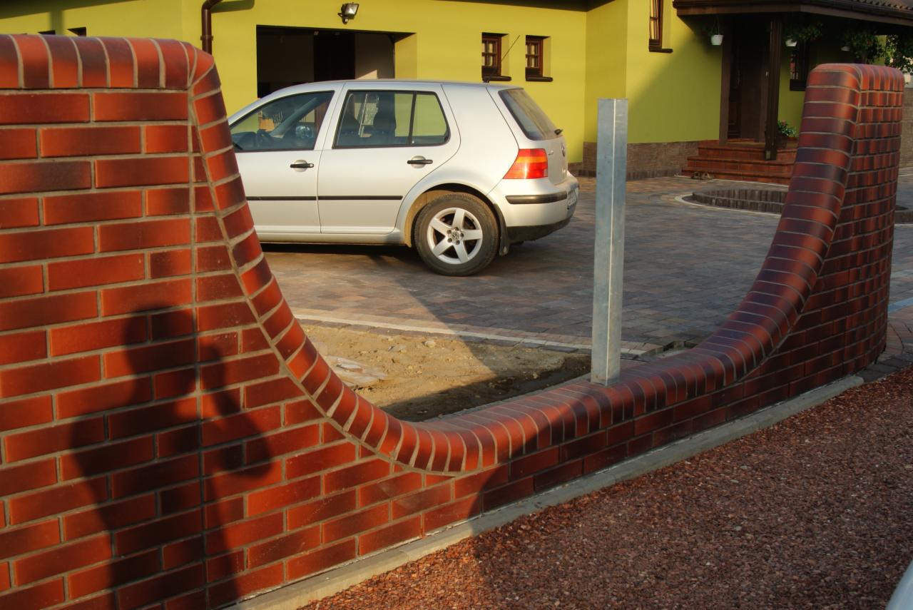 Ceglany, falisty murek ogrodzeniowy z metalowym słupkiem na tle domu i kostki brukowej. Widoczny cień osoby na murku. Ujęcie z dołu.