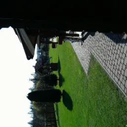 Zadbany ogród z zielonym trawnikiem, ścieżką z kostki brukowej i drewnianą studnią. Widok z góry z lekkim obrotem, ukazujący estetyczne zagospodarowanie przestrzeni.