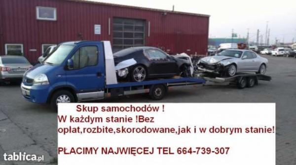 Niebieska laweta z dwoma uszkodzonymi samochodami na platformie, na tle parkingu i budynku. Jeden pojazd jest czarny, drugi srebrny. Widoczny napis o skupie aut.