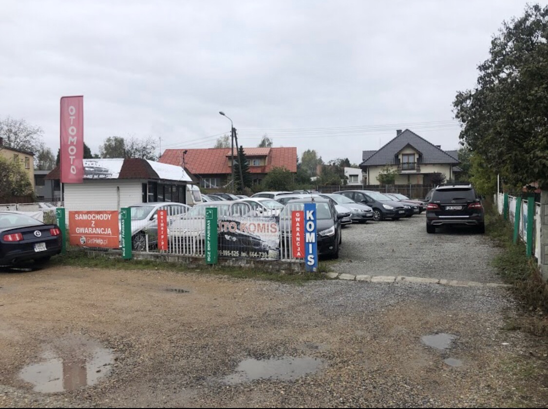 Plac komisu samochodowego z zaparkowanymi pojazdami, widoczne reklamy i budynek biura, pochmurny dzień.