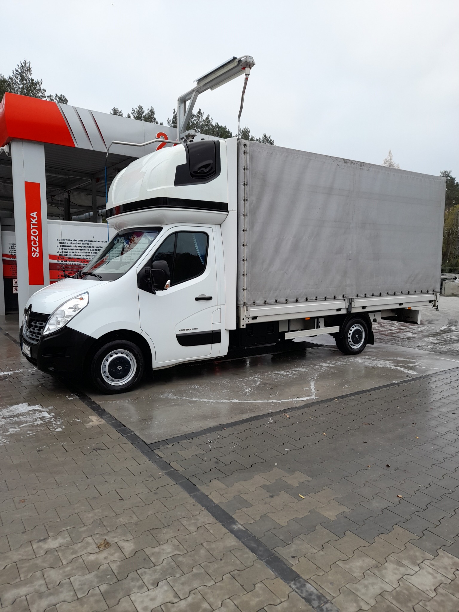 Biały samochód dostawczy z plandeką, po umyciu na myjni automatycznej, z widocznymi śladami wody na kostce brukowej.