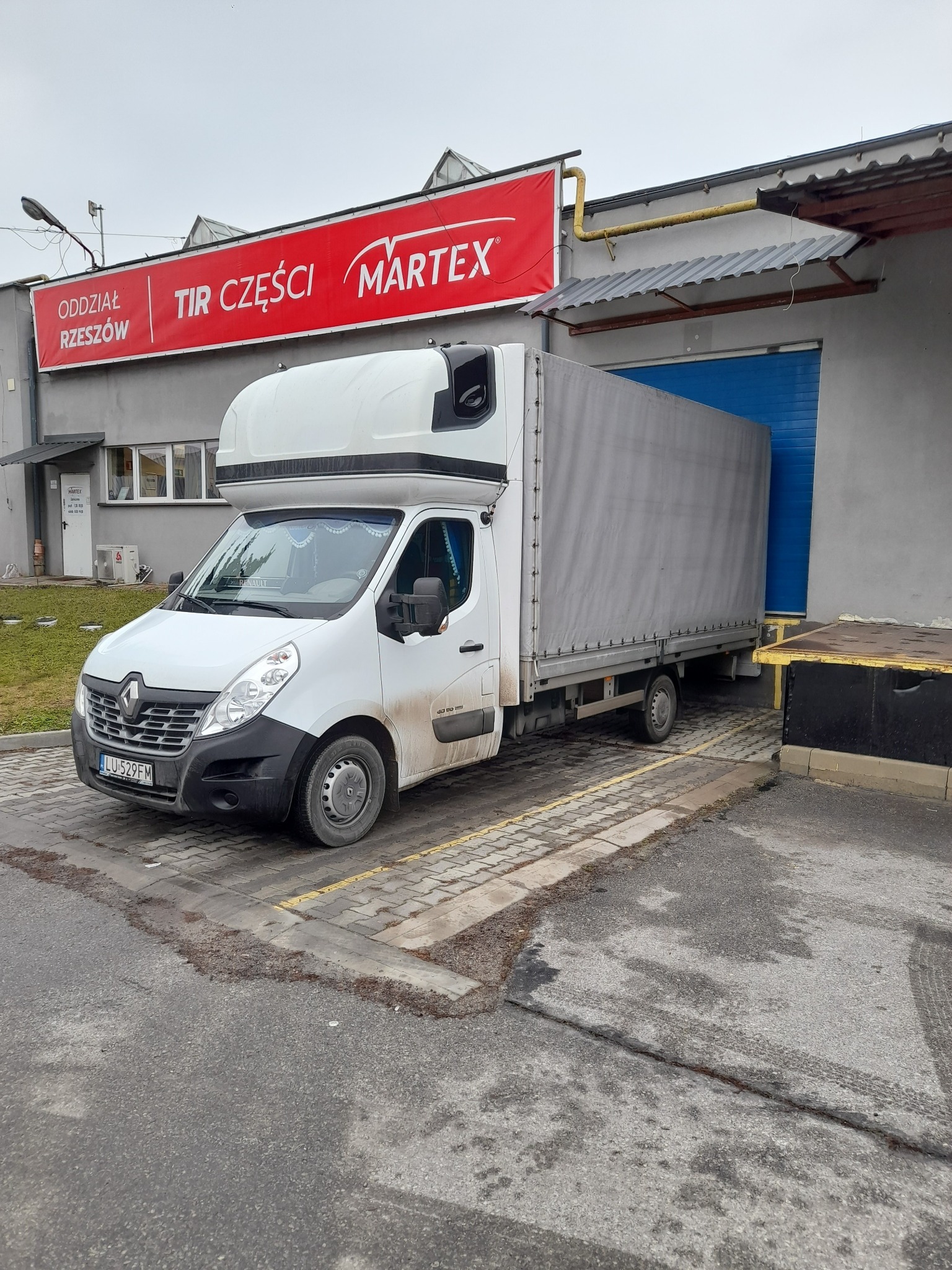 Biały samochód dostawczy Renault Master z plandeką, zaparkowany przed budynkiem firmy Martex w Janowie Lubelskim, z widocznym szyldem 'Oddział Rzeszów TIR Części'.