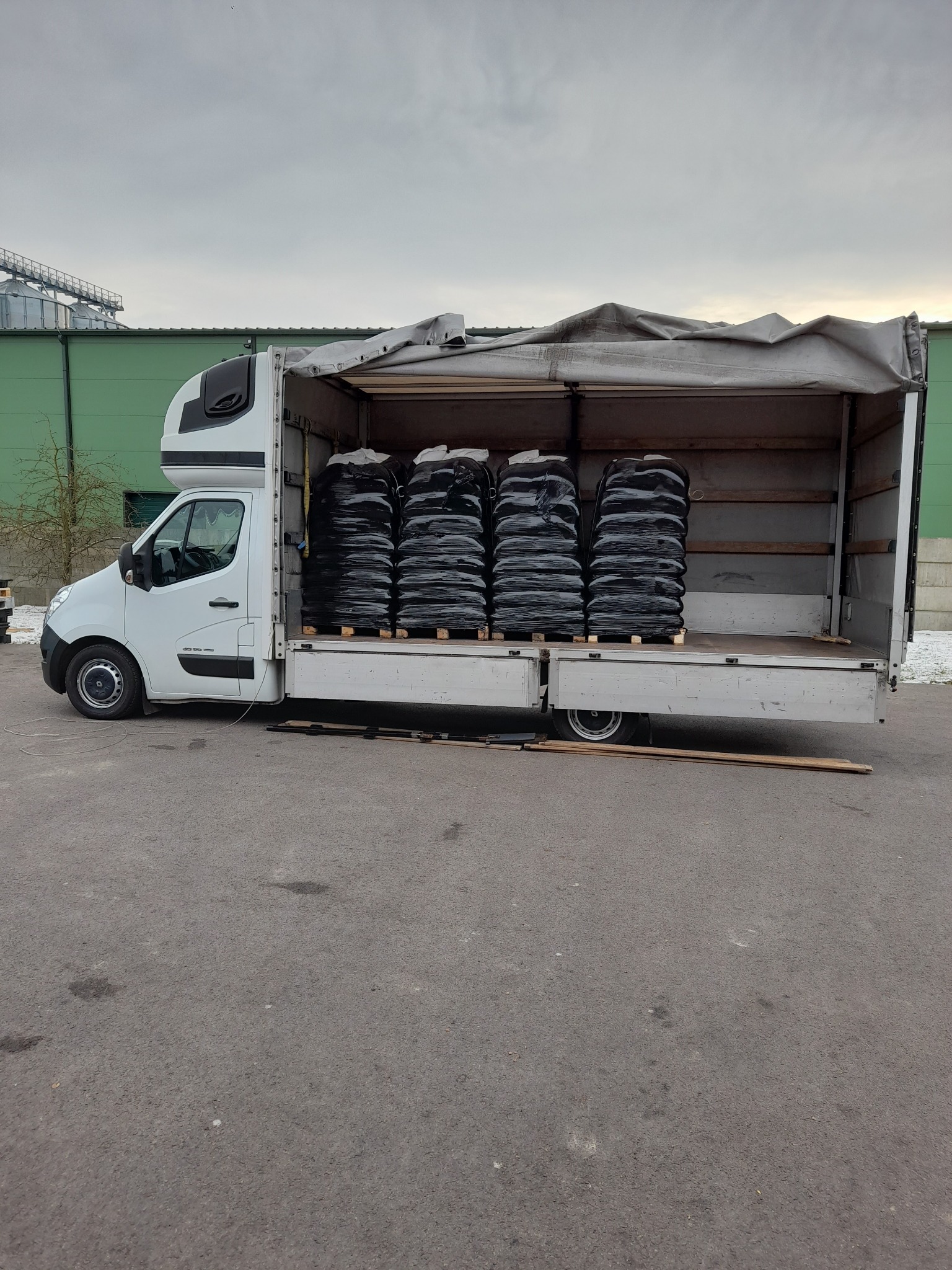 Biały bus z otwartą przestrzenią ładunkową, wypełnioną rzędami czarnych worków na paletach, na tle szarej nawierzchni i zielonego budynku.