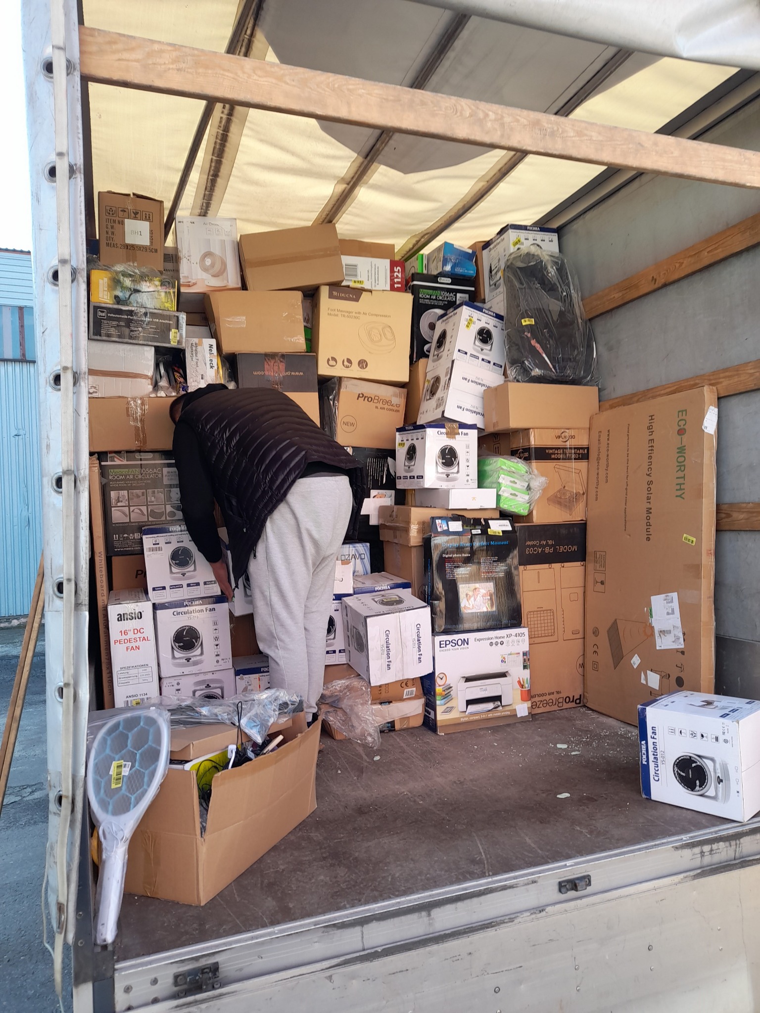 Mężczyzna w kurtce i dresach pochyla się wewnątrz furgonetki wypełnionej kartonami z wentylatorami, drukarkami Epson i modułem solarnym Eco-Worthy, widoczna siatka na owady w kartonie z przodu.
