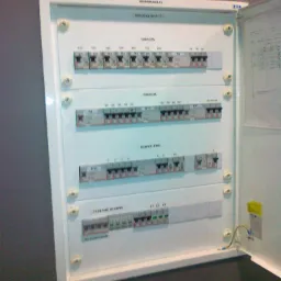 Otwarta skrzynka rozdzielcza z bezpiecznikami, opisana sekcjami: gniazda, oświetlenie, zasilanie główne. Widoczne kable i schemat na wewnętrznej stronie drzwiczek.