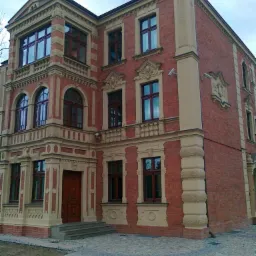 Elewacja odnowionego, trzypiętrowego budynku z czerwonej cegły z beżowymi detalami architektonicznymi i widocznymi kamerami monitoringu.