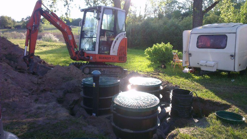 Pomarańczowa mini koparka Schaeff i trzy czarne szamba z zielonymi pokrywami na trawiastym terenie, obok przyczepa kempingowa.