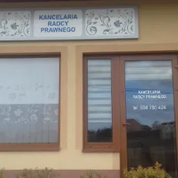 Fasada budynku z szyldem 'Kancelaria Radcy Prawnego' i numerem telefonu, okna z dekoracyjnymi zasłonami, tabliczka informacyjna z danymi radcy prawnego.