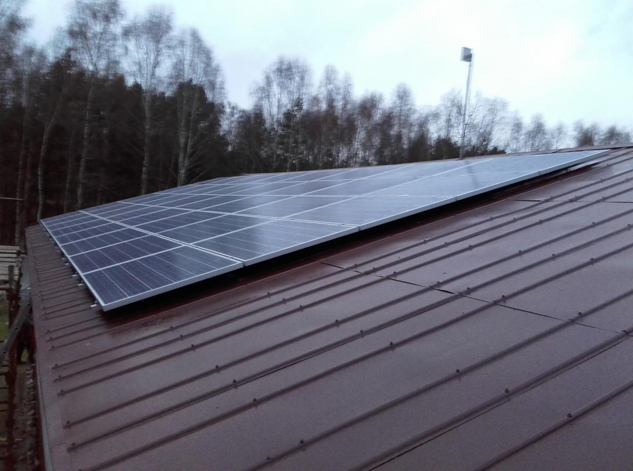 Panel słoneczny na brązowym dachu z blachy trapezowej w pochmurny dzień. W tle las i słup oświetleniowy. Widok z dołu.