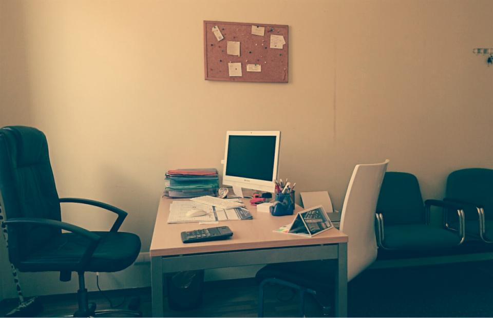 Biuro tłumaczeń: biurko z komputerem, stosem dokumentów, kalkulatorem i przyborami do pisania, tablica korkowa z notatkami na ścianie.