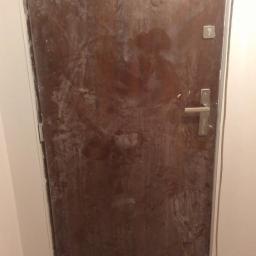 GTech Andrzej Gil - Brudne, brązowe drzwi wejściowe w trakcie prac remontowych, widoczne ślady farby i gipsu. Niechlujne wykończenie, szare płytki podłogowe.