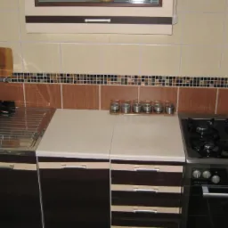 Wykończona kuchnia z brązowymi płytkami i szafkami. Widoczny zlew, płyta gazowa i przyprawy. Całość utrzymana w ciepłej, beżowo-brązowej tonacji.