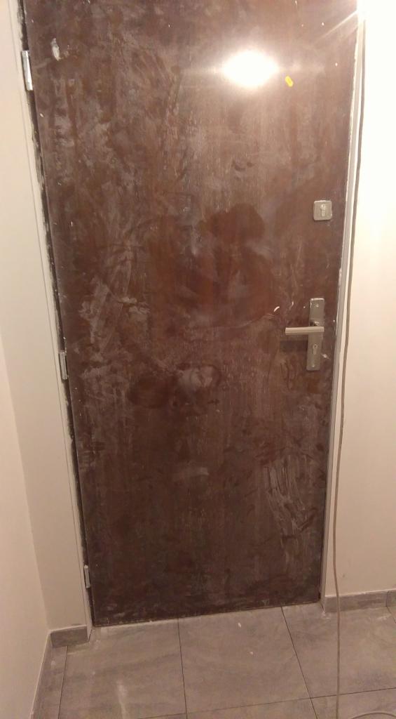 Brudne, brązowe drzwi wejściowe w trakcie prac remontowych, widoczne ślady farby i gipsu. Niechlujne wykończenie, szare płytki podłogowe.