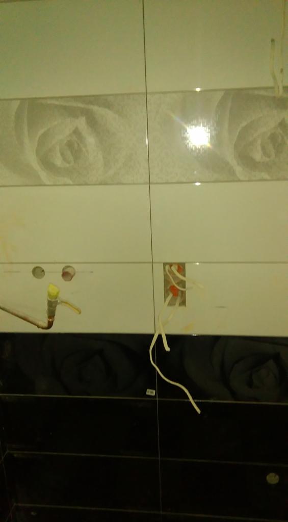 Ściana wyłożona błyszczącymi płytkami w odcieniach bieli i czerni. Widoczne otwory na armaturę i wystające przewody elektryczne, sugerujące etap prac wykończeniowych.