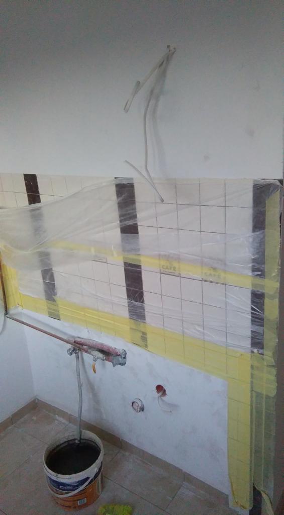 Przygotowanie do prac w łazience: zabezpieczone folią i taśmą płytki, wiadro z farbą i narzędzia malarskie. Widoczne gniazdka elektryczne.