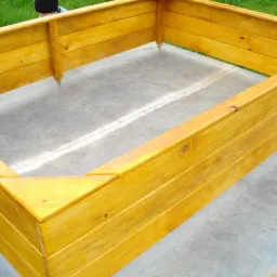 Drewniana, żółta obudowa rabaty warzywnej na betonowym podłożu. Konstrukcja z desek, widoczne łączenia i faktura drewna. Ujęcie z góry, częściowo widoczne trawa.