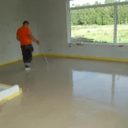 Aplikacja wylewki samopoziomującej w jasnym pomieszczeniu z dużym oknem. Pracownik wyrównuje powierzchnię długim narzędziem. Żółta taśma wzdłuż ścian.