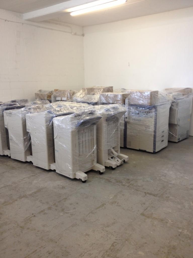 Magazyn z używanymi kserokopiarkami A3 do A0, zabezpieczonymi folią stretch, ustawionymi rzędami na betonowej podłodze pod jarzeniówkami.